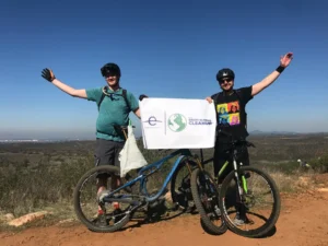 Two men with bicycles stand on a dirt path at the top of a hill, holding up a sign that reads "The Great Global Cleanup"