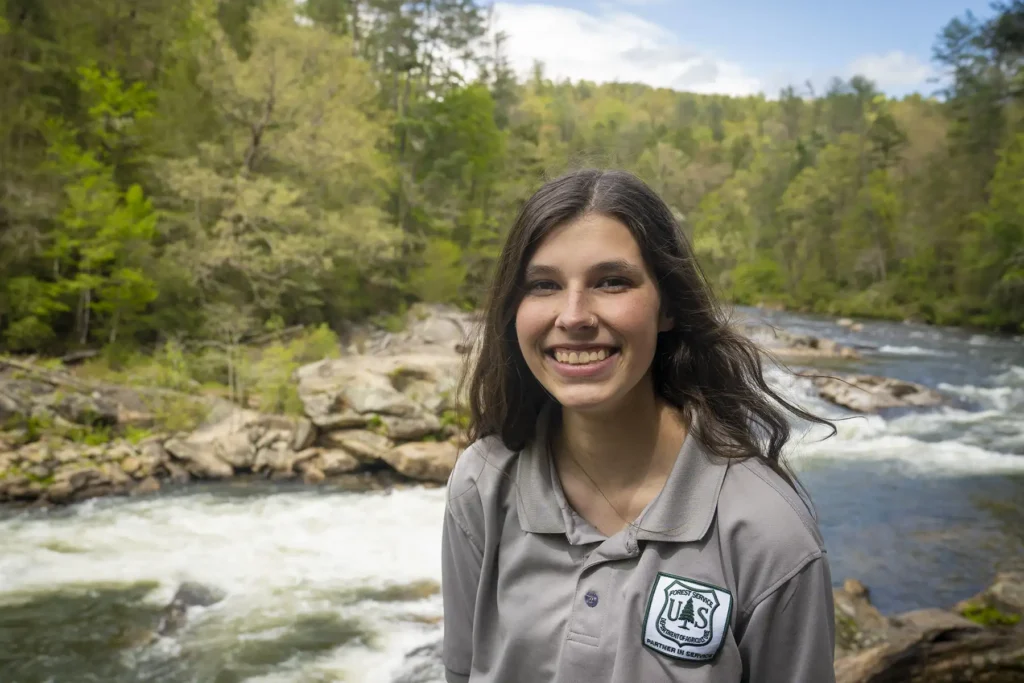 A young woman from the US Forest Service smiling in front of a river surrounded by trees.